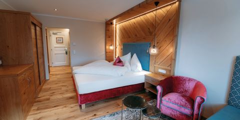 Modernes Hotelzimmer mit Doppelbett, Holzmöbeln, rosa Sessel, kleinen Tischen und warmer Beleuchtung auf einem hölzernen Kopfteil.
