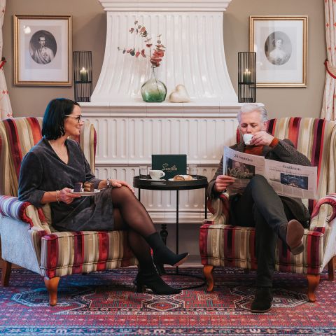 Ein älteres Paar sitzt in Sesseln am Kamin, plaudert und trinkt Kaffee; der Mann liest eine Zeitung. Der Raum ist gemütlich eingerichtet.