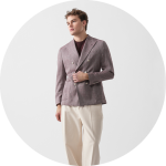 Mann, der einen lilafarbenen Zweireiher über einem dunklen Hemd und einer cremefarbenen Hose trägt, steht vor einem hellen, runden Hintergrund.