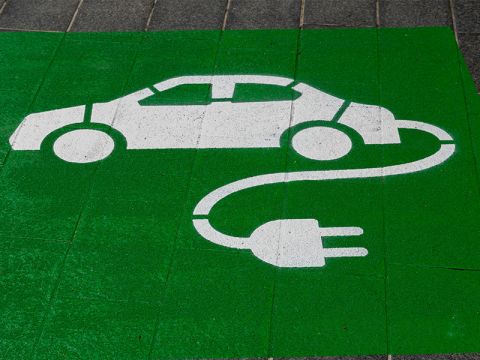 Weißes Elektroauto und Steckersymbol auf grünem Pflaster, das auf eine Ladestation für Elektrofahrzeuge hinweist.