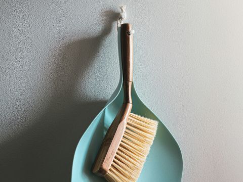 Ein Pinsel mit Holzstiel ruht in einer hellblauen Kehrschaufel, beide hängen an einer weißen Schnur an einer strukturierten Wand.