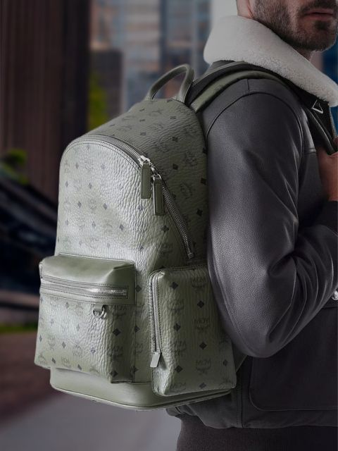 Eine Person in einer schwarzen Jacke trägt einen großen, strukturierten olivgrünen MCM-Rucksack mit Reißverschlusstaschen und steht im Freien in einer städtischen Umgebung.