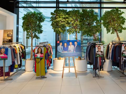 Kleiderständer und große Topfpflanzen umgeben eine Auslage mit Postern von Menschen und einem zentralen Schild in einem hellen, modernen Innenraum.