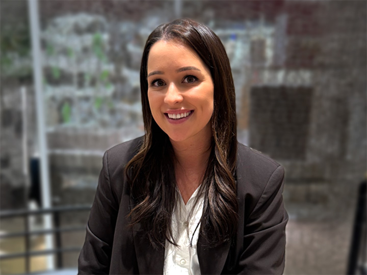 Eine Frau mit langen dunklen Haaren in einem schwarzen Blazer und weißem Hemd lächelt, während sie in einem Haus sitzt, mit einem unscharfen Hintergrund.
