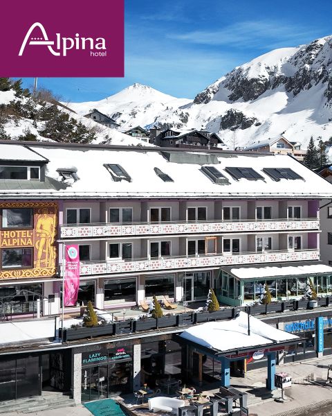 Hotel Alpina mit schneebedecktem Dach und Bergen im Hintergrund, an einem sonnigen Wintertag, mit Geschäften und Sitzgelegenheiten im Freien darunter.