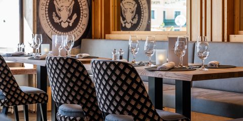 Eleganter, mit Weingläsern, Servietten und Kerzen gedeckter Restauranttisch; gemusterte Stühle und Präsidentensiegel im Hintergrund.