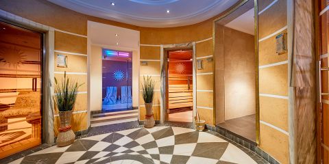 Runder Wellnessbereich mit gemustertem Fliesenboden, warm getönten Wänden, Topfpflanzen und Türen, die zu Saunaräumen mit sanfter Farbbeleuchtung führen.