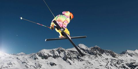 Ein Skifahrer in farbenfroher Ausrüstung vollführt einen Sprungtrick hoch über verschneiten Berggipfeln unter einem klaren, blauen Himmel.