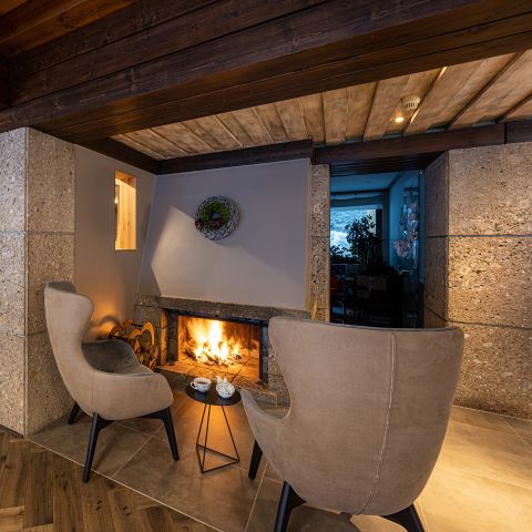 In einem gemütlichen Raum mit Holzdecke und Steinwänden stehen zwei moderne Sessel vor einem beleuchteten Kamin, dazwischen ein kleiner runder Tisch mit Tassen und einer Teekanne.