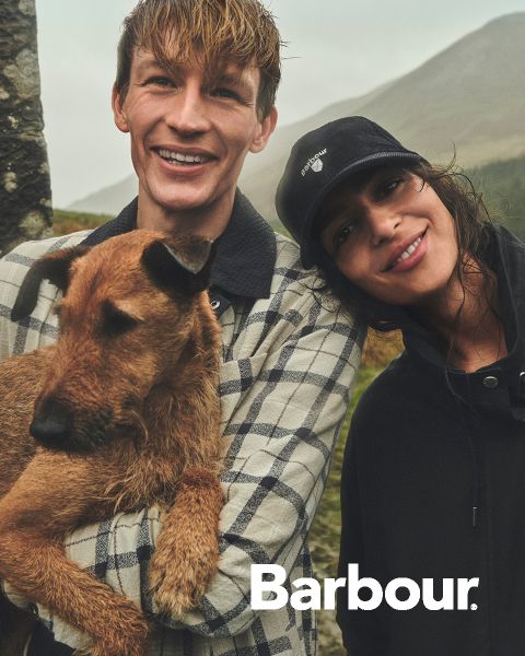 Ein lächelnder Mann mit einem braunen Hund steht neben einer lächelnden Frau mit einer Mütze, beide im Freien mit Hügeln im Hintergrund. Das Barbour-Logo ist sichtbar.