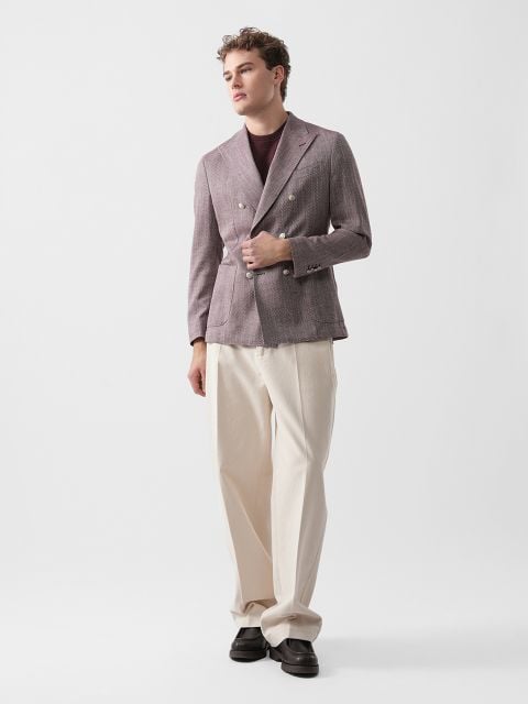 Ein Mann, der einen malvenfarbenen Zweireiher, ein dunkles Hemd und eine weite cremefarbene Hose trägt, steht vor einem schlichten weißen Hintergrund.