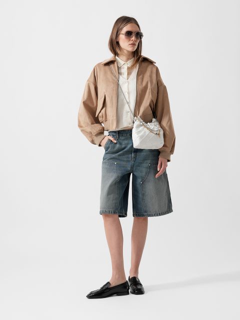 Eine Frau, die eine Sonnenbrille, eine hellbraune Jacke, ein weißes Hemd, lange Jeans-Shorts, schwarze Halbschuhe und eine weiße Crossbody-Tasche trägt, steht vor einem schlichten Hintergrund.