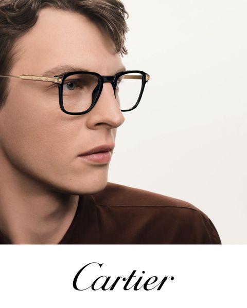 Ein Mann mit einer schwarz-goldenen Brille blickt zur Seite. Er trägt ein braunes Hemd. Am unteren Rand steht das Wort "Cartier".