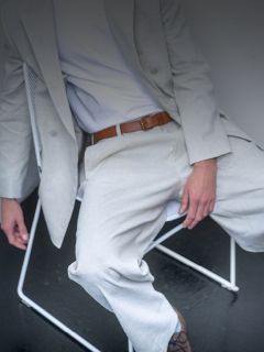 Eine Person in einem hellgrauen Anzug und weißem Hemd sitzt auf einem weißen Stuhl, ein brauner Gürtel ist zu sehen und ihr Gesicht ist nicht zu sehen.