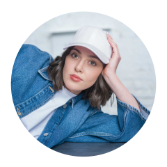 Frau mit weißer Mütze und Jeansjacke, die sich auf ihren Arm stützt und mit neutralem Blick in die Kamera schaut.