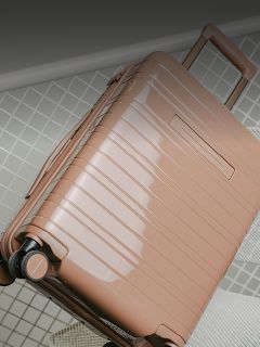 Ein glänzender, hellbrauner Hartschalenkoffer mit Rollen und einem verlängerten Griff steht auf einem Fliesenboden.