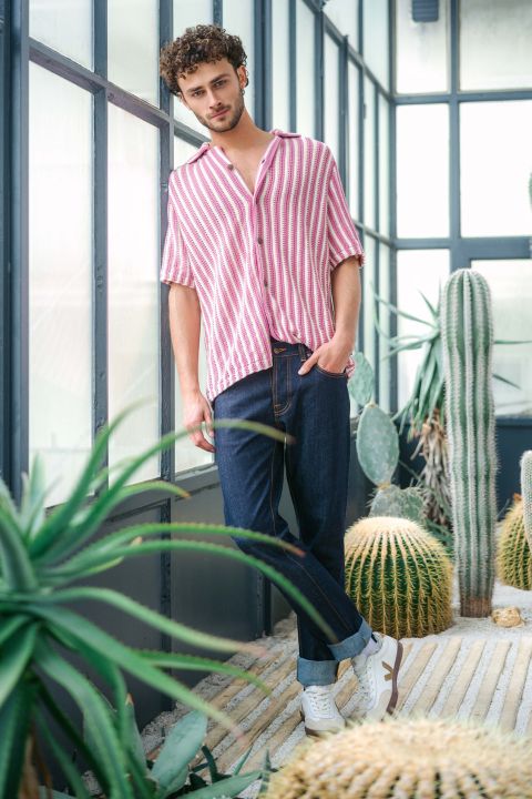 Ein Mann in einem rosa-weiß gestreiften Hemd und dunklen Jeans steht in einem hellen, gläsernen Raum zwischen Kakteen und schaut in die Kamera.