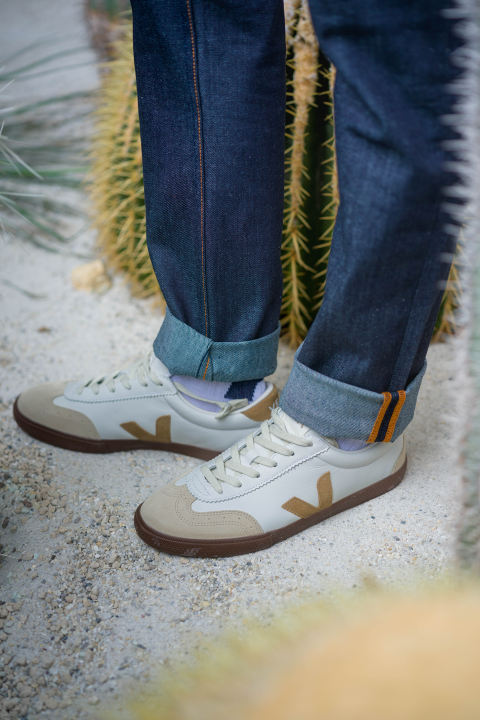 Eine Person in hochgekrempelten Jeans und weißen Sneakers mit hellbraunen Akzenten, die auf Kies neben einem großen Kaktus steht.