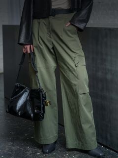 Person, die eine olivgrüne Cargohose, schwarze Schuhe und eine schwarze Jacke trägt und eine glänzende schwarze Handtasche an ihrer Seite hält.