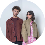 Zwei Personen stehen nebeneinander; die eine trägt eine braune Jacke, die andere eine Sonnenbrille und eine hellbraune Jacke über einem rosa Kleid mit kreisförmigen Mustern.