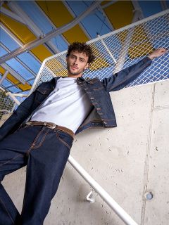 Ein Mann in weißem T-Shirt, dunkler Jeansjacke und Jeans steht an einem Geländer, das an eine Betonmauer und einen Zaun gelehnt ist, darüber eine gelbe Markise.