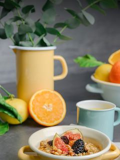 Eine Schale mit Müsli, Feigen und Beeren, ein gelber Krug mit grünen Zweigen, Zitrusfrüchte und eine hellblaue Tasse auf einem grauen Tisch.