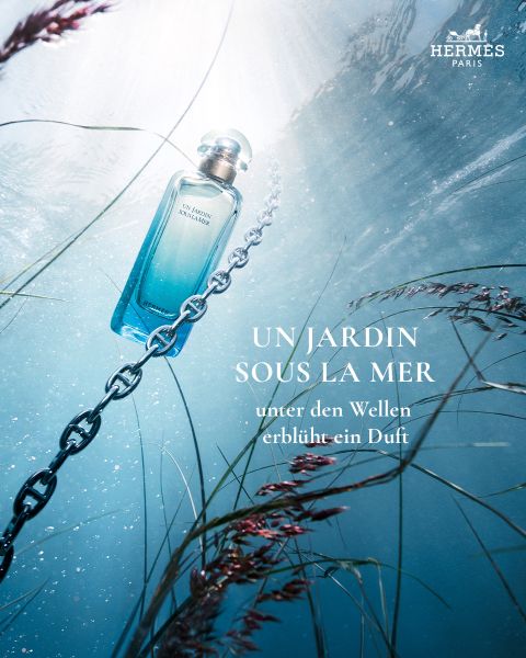 Ein blauer Parfümflakon ist unter Wasser an einer Kette befestigt, umgeben von Pflanzen, durch die das Sonnenlicht fällt und das Branding von Hermès Paris sichtbar ist.