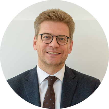 Lächelnder Mann mit kurzem, hellbraunem Haar und Brille, der einen dunklen Anzug, ein weißes Hemd und eine gemusterte Krawatte trägt, vor einem hellen Hintergrund.