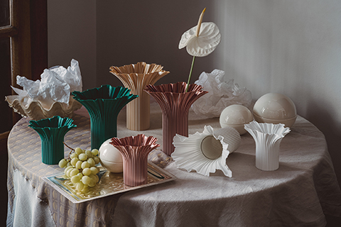 Auf einem Tisch stehen bunte geriffelte Vasen, weiße Zierkugeln, ein Teller mit Weintrauben und zerknülltes weißes Papier in sanftem Tageslicht.