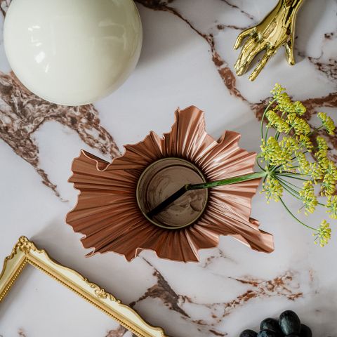 Eine bronzene, geriffelte Vase mit gelben Blumen steht auf einer Marmorfläche, umgeben von einer goldenen Hand, einem runden weißen Objekt, einem Bilderrahmen und Weintrauben.