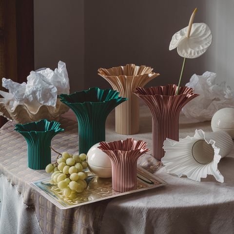 Bunte geriffelte Vasen, weiße skulpturale Objekte, Weintrauben und eine weiße Blume sind auf einem drapierten Tisch mit einem strukturierten Hintergrund angeordnet.