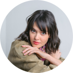 Frau mit dunklem, schulterlangem Haar und Pony, in hellbrauner Jacke, lehnt sich mit verschränkten Armen vor und blickt in die Kamera.