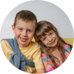 Zwei lächelnde Kinder, ein Junge in einem gelben Hemd und ein Mädchen in einem gestreiften Hemd, sitzen dicht beieinander auf einer gelben Couch vor einem hellen Hintergrund.