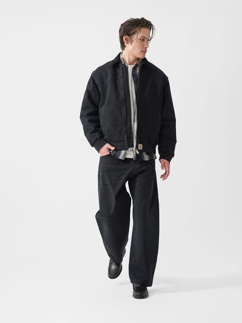 Eine Person, die eine schwarze Jacke, eine schwarze Hose und schwarze Schuhe trägt, geht selbstbewusst vor einem schlichten weißen Hintergrund.