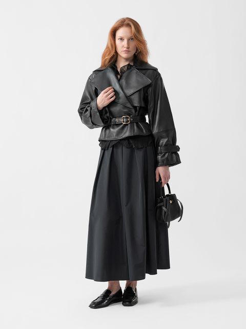 Eine Frau mit langen roten Haaren trägt eine schwarze Lederjacke, einen langen schwarzen Rock, schwarze Schuhe und hält eine kleine schwarze Handtasche vor einem schlichten Hintergrund.