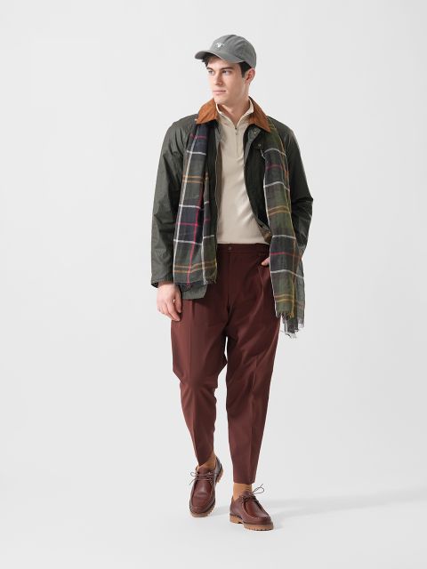 Ein Mann mit grauer Mütze, grüner Jacke, kariertem Schal, beigem Hemd, brauner Hose und braunen Schuhen steht vor einem schlichten weißen Hintergrund.