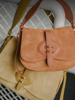 Zwei Handtaschen aus Wildleder, eine hellbraun und eine beige, mit Lederriemen und goldenen Verschlüssen, die auf einem Hintergrund aus weißem Metallgitter hängen.