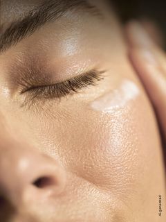 Eine Nahaufnahme des Gesichts einer Person mit geschlossenen Augen, die glatte, strahlende Haut und einen Hauch von Creme unter dem Auge zeigt.