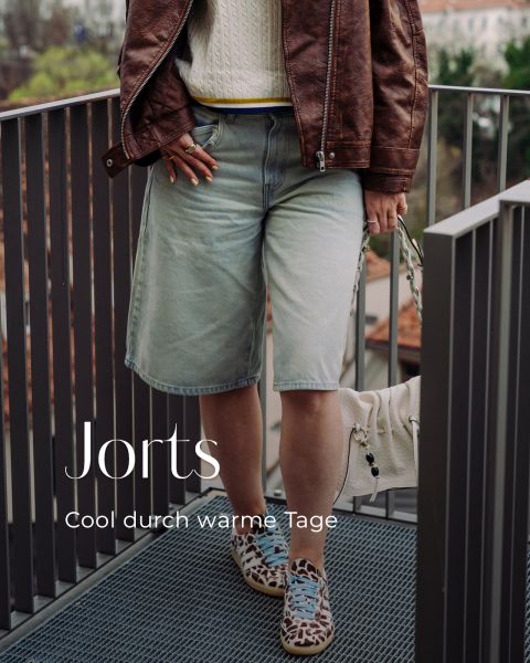 Person in brauner Lederjacke, weißem Strickpullover, Jeans-Jorts und gemusterten Turnschuhen, die auf einem Balkon steht. Der Text lautet: "Jorts Cool durch warme Tage.