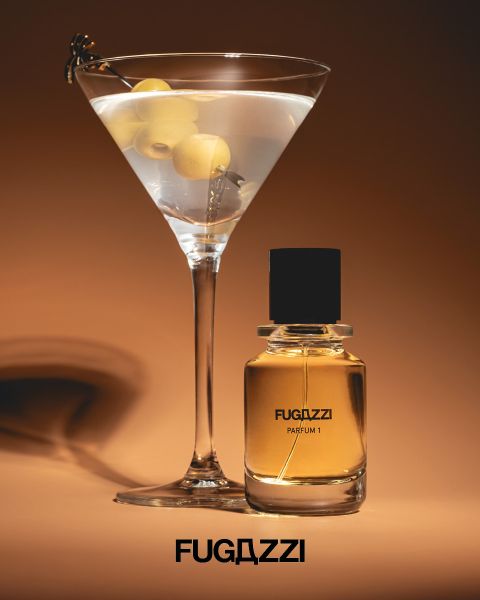 Ein Martini-Glas mit Oliven neben einem Flakon von Fugazzi Parfum I auf braunem Hintergrund, darunter das Fugazzi-Logo.