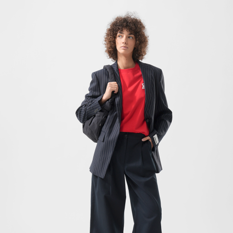 Frau mit lockigem Haar, die einen Nadelstreifenblazer, ein rotes Hemd und eine Hose mit weitem Bein trägt und eine schwarze Tasche mit sich führt, steht vor einem schlichten weißen Hintergrund.