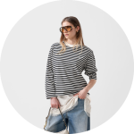 Frau mit einer großen Sonnenbrille, einem schwarz-weiß gestreiften Pullover, blauen Jeans und einer weißen Handtasche in der Hand vor einem hellen Hintergrund.