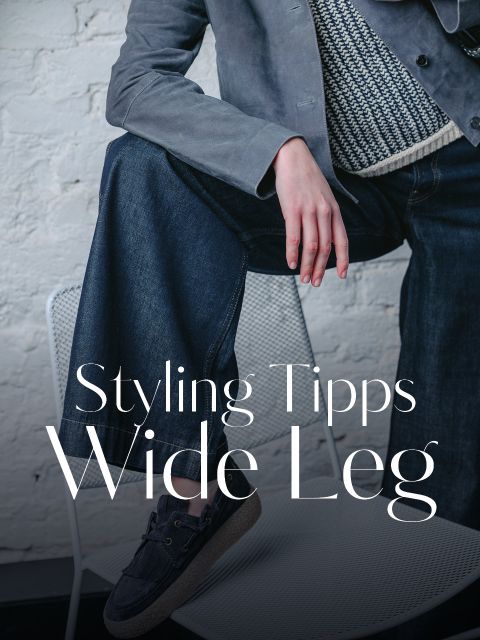 Eine Person, die eine Jeans mit weitem Bein, einen Strickpullover und eine graue Jacke trägt, sitzt auf einem weißen Stuhl. Der Text lautet: „Styling-Tipps für weite Hosenbeine.“.