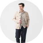 Mann mit beiger Jacke über einem gestreiften Hemd und dunkler Hose, stehend und zur Seite blickend vor einem hellgrauen Hintergrund.