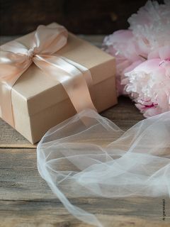 Geschenkkarton mit zartrosa Schleife neben zartrosa Pfingstrosen und weißem Stoff auf einer Holzoberfläche.