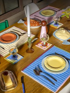 Ein hölzerner Esstisch mit bunten Tellern, gesteppten Tischsets, Gläsern, Besteck und Kerzen in einem hellen, eklektischen Arrangement.