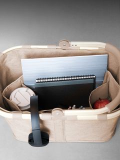 Ein beiger Korb enthält einen Laptop, ein Notizbuch, Stifte, einen weißen Reisebecher und einen roten Apfel, von oben betrachtet.