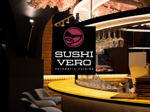 Eine moderne Sushi-Bar mit einem runden, gelb beleuchteten Tresen, hängenden Gläsern und einem "Sushi Vero Authentic Cuisine"-Schild mit einem Fisch-Logo.