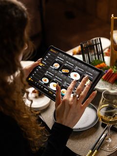 Eine Person blättert mit einem Tablet in einer digitalen Restaurantkarte an einem mit Essen, Wein und Stäbchen gedeckten Tisch.