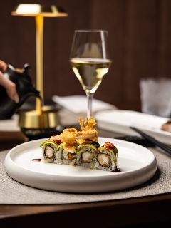 Ein Teller mit Sushi-Rollen, belegt mit Avocado und knuspriger Garnierung, serviert mit einem Glas Weißwein an einem gedeckten Tisch.